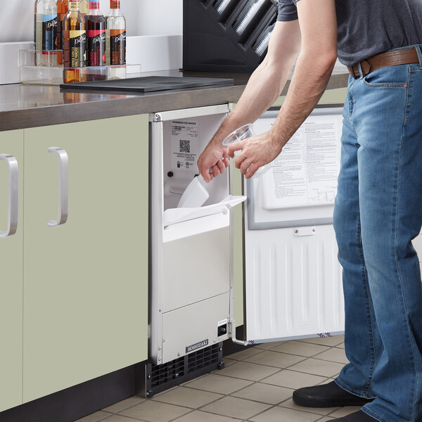 A man putting a plastic cup of ice into a white rectangular undercounter refrigerator.