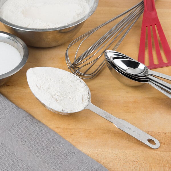 A Vollrath stainless steel measuring scoop filled with flour on a table with measuring spoons and a bowl of flour.