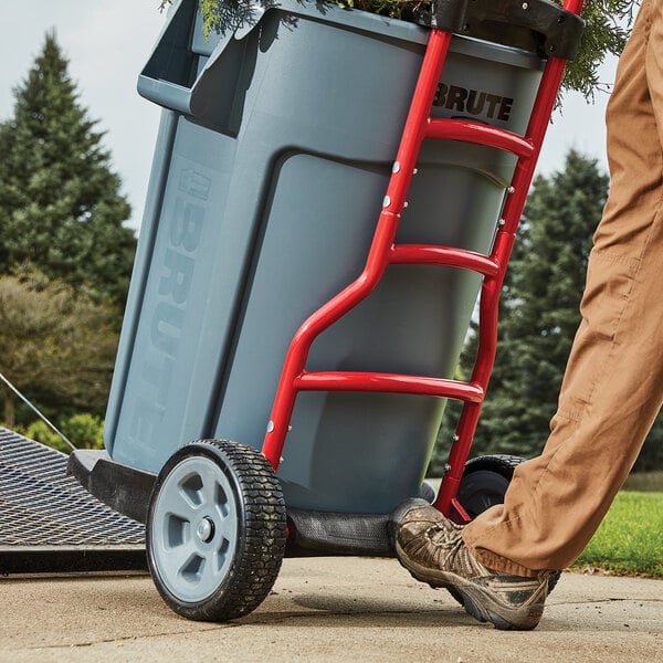 A man pushing a large grey Rubbermaid trash can on a dolly.