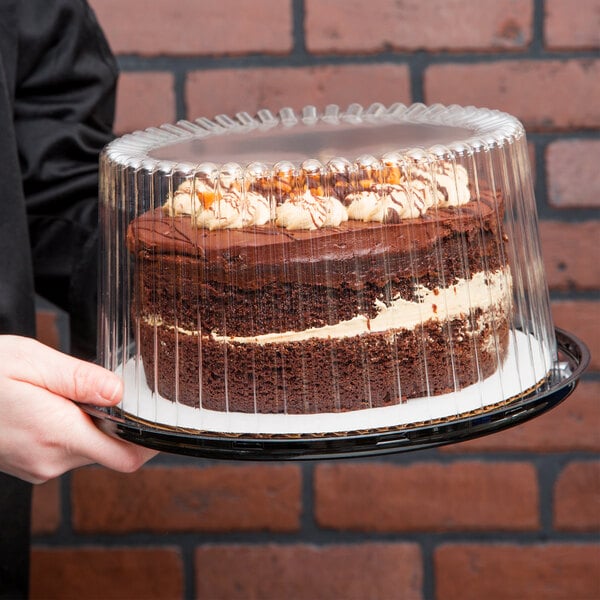 A person holding a chocolate cake in a D&W Fine Pack clear plastic cake container.