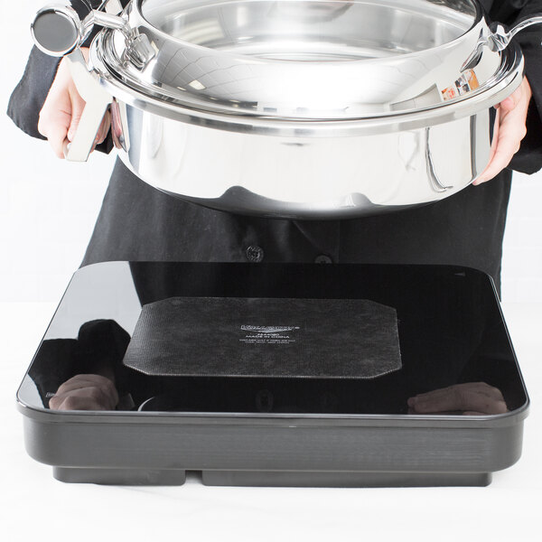 A person holding a silver pot over a black surface with a black Vollrath non-slip silicone chafer mat inside.