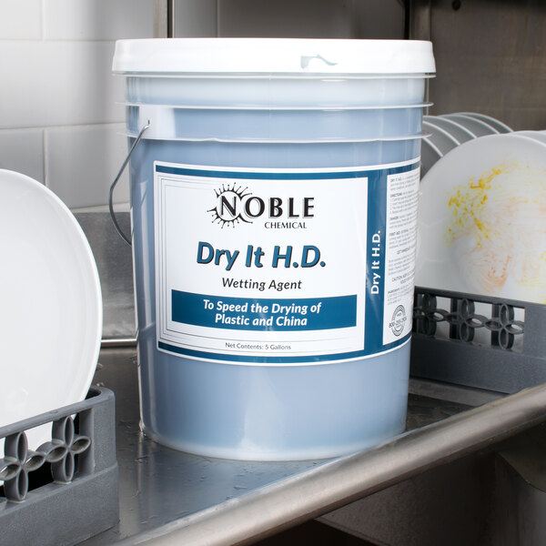 A 5 gallon bucket of Noble Chemical Dry It HD+ sitting on a counter in a professional kitchen.