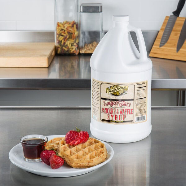A white jug of Golden Barrel sugar free pancake and waffle syrup on a table next to a plate of waffles with strawberries.