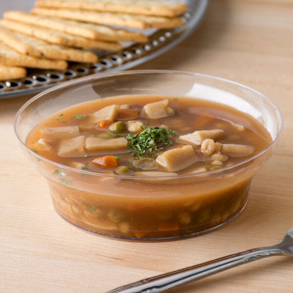 A clear Fineline disposable plastic bowl of soup with crackers on a table.