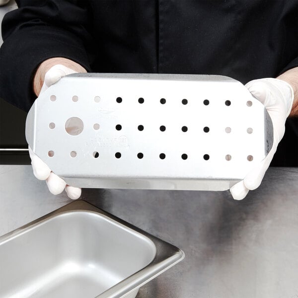 A person in gloves holding a stainless steel Vollrath false bottom with holes.