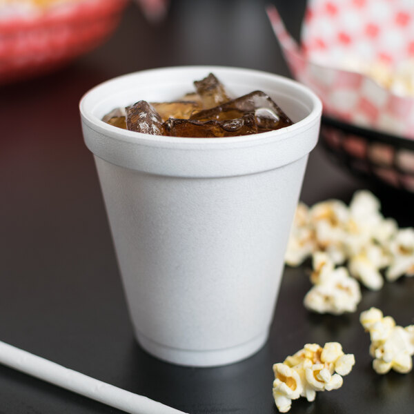 A Dart white foam cup filled with soda and ice with popcorn on the table.