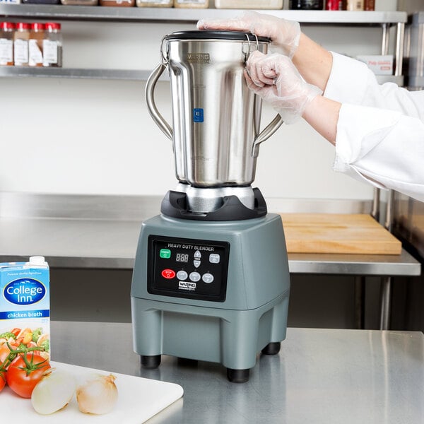 A person in a white coat and gloves using a Waring stainless steel food blender on a counter in a professional kitchen.