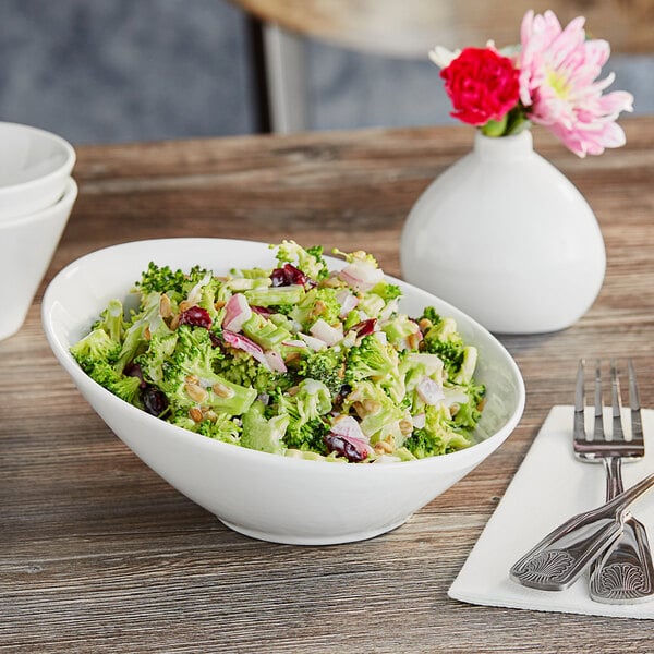A white Acopa porcelain bowl filled with broccoli salad on a table with a vase of flowers.