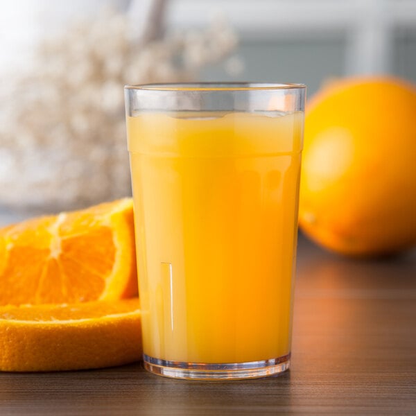 A Carlisle clear plastic tumbler filled with orange juice next to oranges.