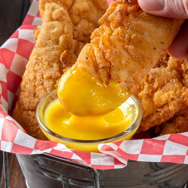A hand dips a Ken's Foods Honey Mustard sauce-covered chicken strip into a bowl of yellow liquid.