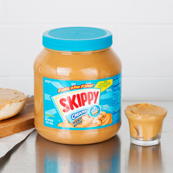 A jar of Skippy Creamy Peanut Butter on a table next to a spoon and a glass of peanut butter.