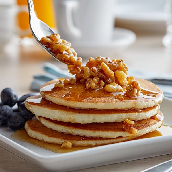 A spoon pouring J. Hungerford Smith Maple Walnut Dessert Topping onto a stack of pancakes.