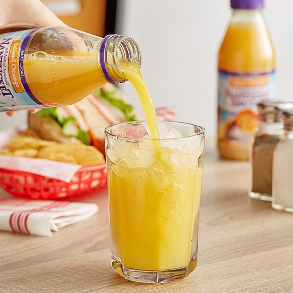 A hand pouring Nantucket Nectars Premium Orange Juice into a glass with ice.