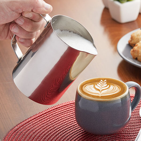 A hand holding a Choice stainless steel pitcher pouring milk into a cup of coffee.