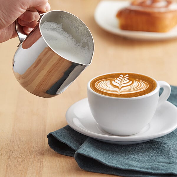 A person using a Choice stainless steel frothing pitcher to pour milk into a cup of coffee.