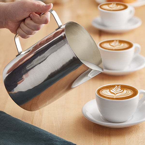 A person pouring milk from a Choice stainless steel frothing pitcher into a cup of coffee with a leaf design in the foam.
