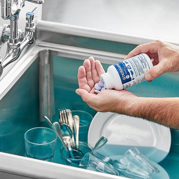 A person washing dishes in a sink with a bottle of Edwards-Councilor S150E48 Steramine Sanitizer Tablets on the counter.