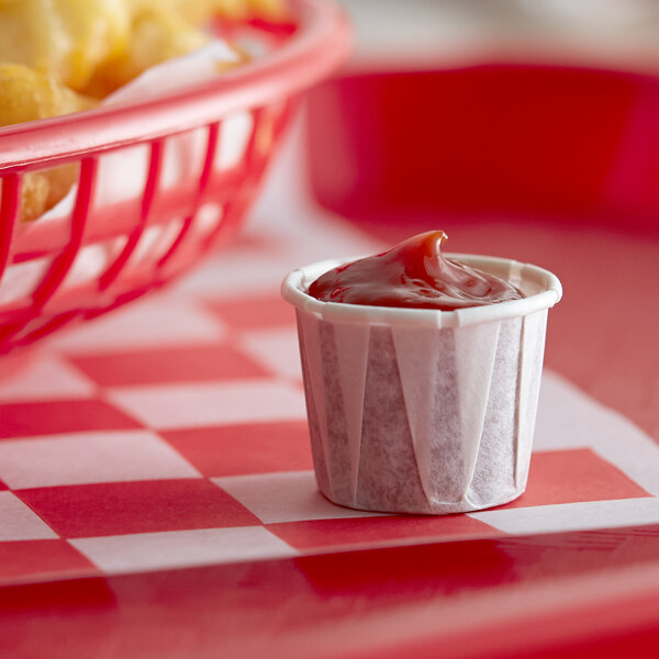 A small Genpak paper souffle cup of ketchup on a table with a basket of fries.