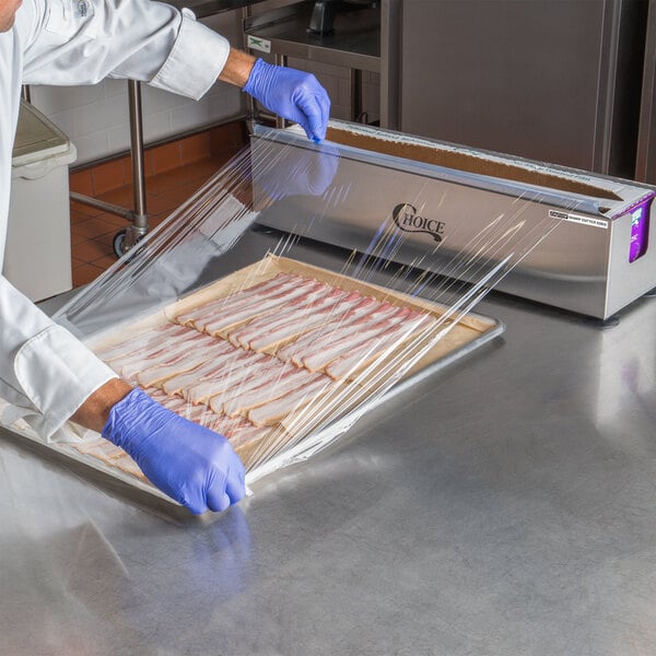 A person in a blue coat uses the Choice stainless steel film and foil cutter to cut plastic wrap over a tray of bacon.