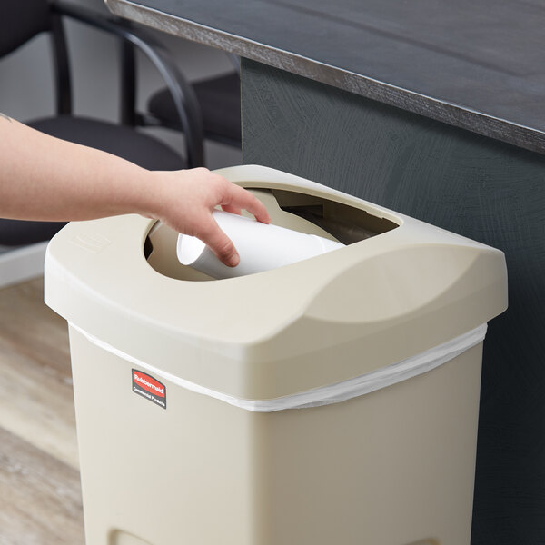 A hand putting a paper cup into a white Rubbermaid Untouchable trash can.