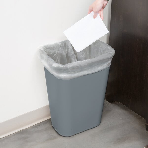 A hand throwing a piece of paper into a Rubbermaid grey rectangular trash can.