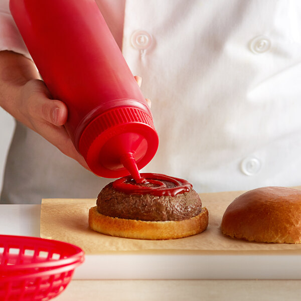 A hand using a Vollrath red wide mouth squeeze bottle to put ketchup on a hamburger.