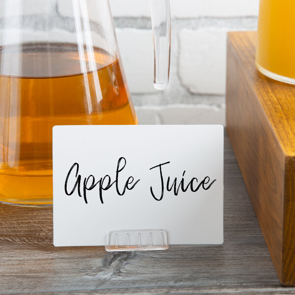 A mini white chalk card with black text next to a glass of yellow apple juice.