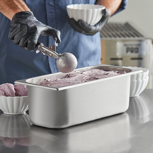 A man in gloves using a scoop to fill a white bowl with ice cream from a stainless steel pan.