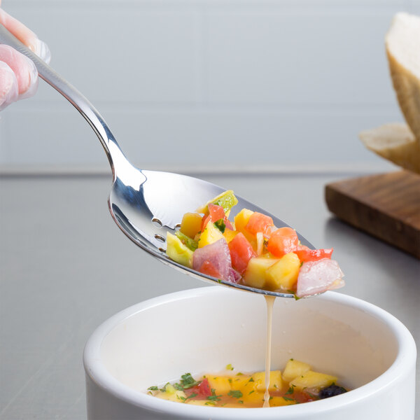 A hand using a Mercer Culinary stainless steel perforated bowl plating spoon to scoop fruit from a bowl.