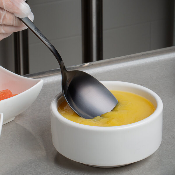 A hand holding a Mercer Culinary black plating spoon in a bowl of yellow liquid.