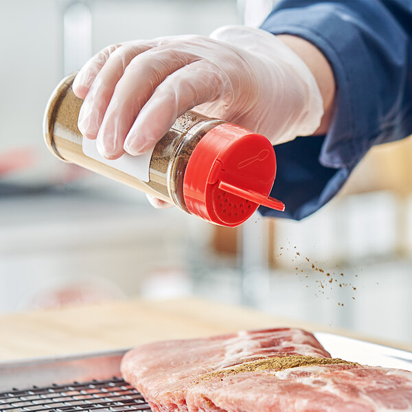 A hand in a glove holding a 53/485 Round Plastic Spice Container pouring seasoning on meat.