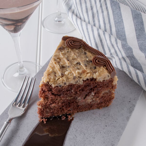 A slice of Pellman German Chocolate Cake with chocolate frosting and chocolate chips on top on a plate with a fork and a glass of liquid.