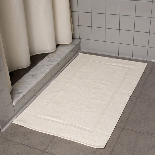 An Oxford Vicenza Avorio white bath mat with a dobby border on a tile floor.