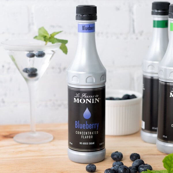 A bottle of Monin Blueberry Concentrated Flavor on a table next to a white container of blueberries.