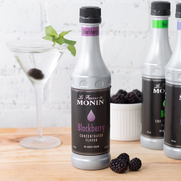 A close-up of a Monin Blackberry Concentrated Flavor bottle on a table next to a martini glass.