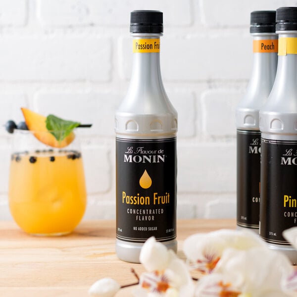A close-up of a bottle of Monin Passion Fruit Concentrated Flavor next to a glass of orange juice on a table.