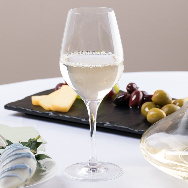 A Stolzle Exquisit Chardonnay wine glass filled with white wine on a table with a plate of olives and cheese.
