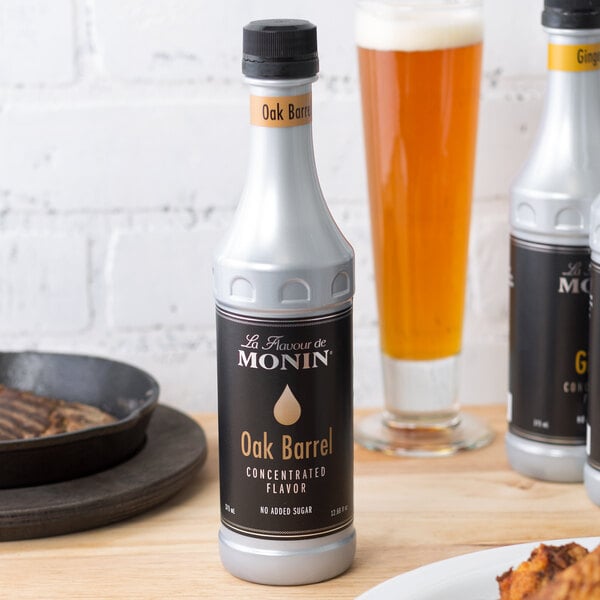 A black Monin Oak Barrel Concentrated Flavor bottle with gold text on a table next to a plate of food and a glass of orange liquid.