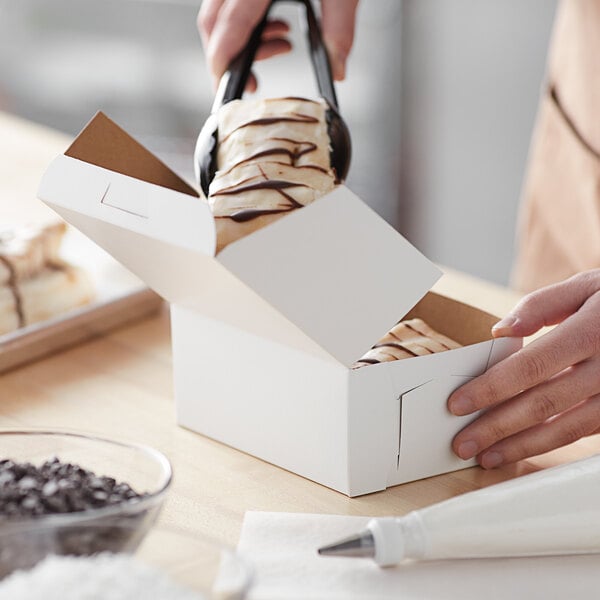 A hand putting a piece of food into a white Baker's Mark bakery box.