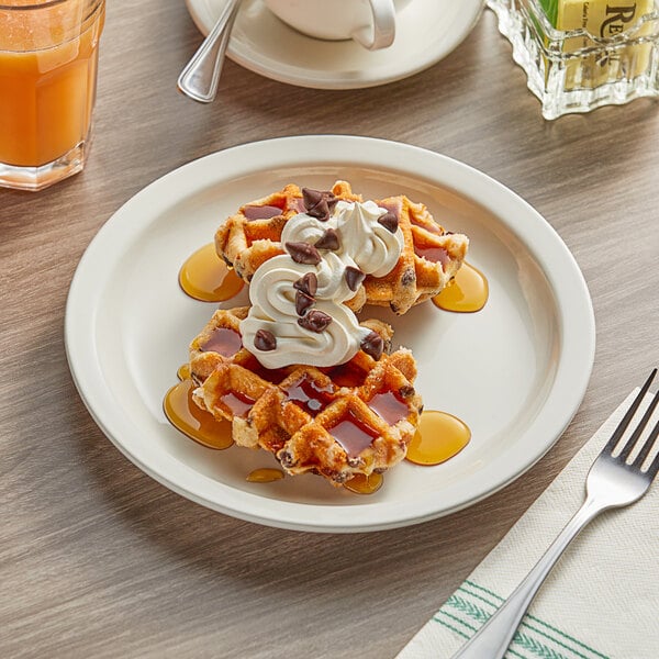 An Acopa narrow rim stoneware plate with waffles, whipped cream, and syrup on a table with a fork and spoon.