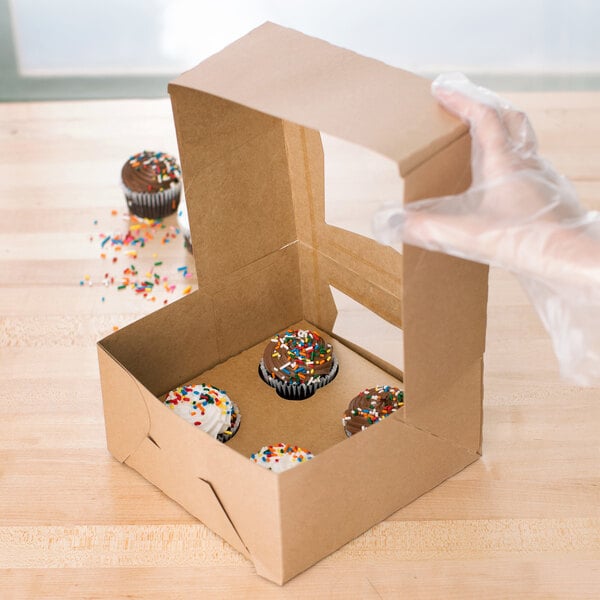 A hand holding a Baker's Mark kraft cupcake box with a chocolate cupcake and sprinkles inside.