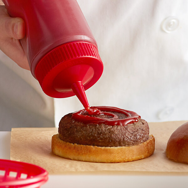A hand using a Vollrath Color-Mate squeeze bottle with a red cap to put ketchup on a hamburger.