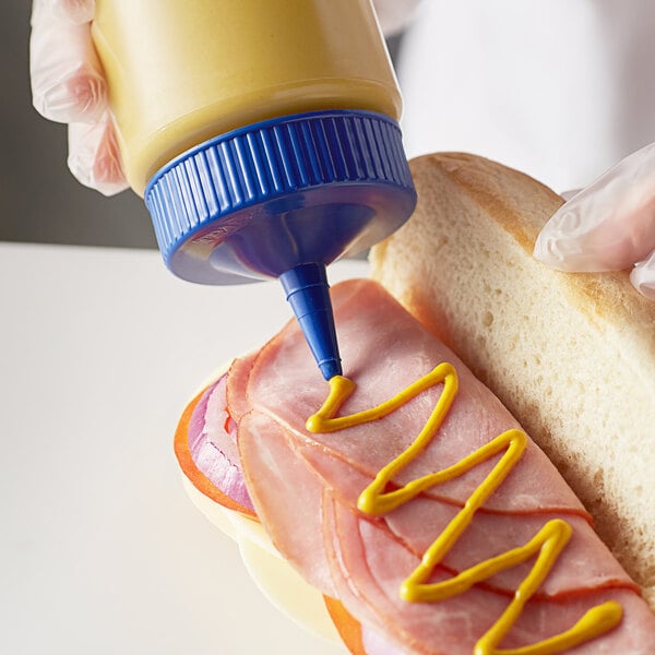 A person holding a Vollrath Color-Mate squeeze bottle with blue cap and putting mustard on a sandwich.