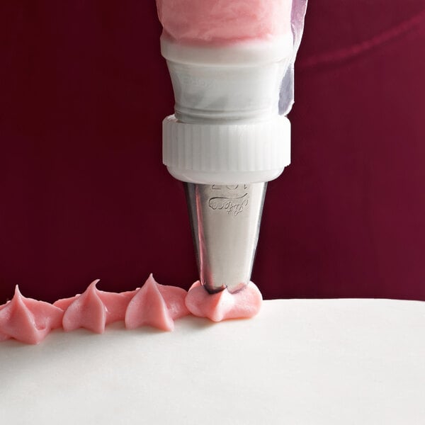 A person using an Ateco Drop Flower piping tip in a white pastry bag to pipe pink frosting on the edge of a cake.