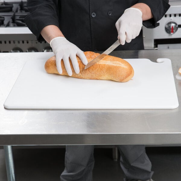 A person in a white shirt and gloves cutting a loaf of bread with a San Jamar white cutting board and a knife.