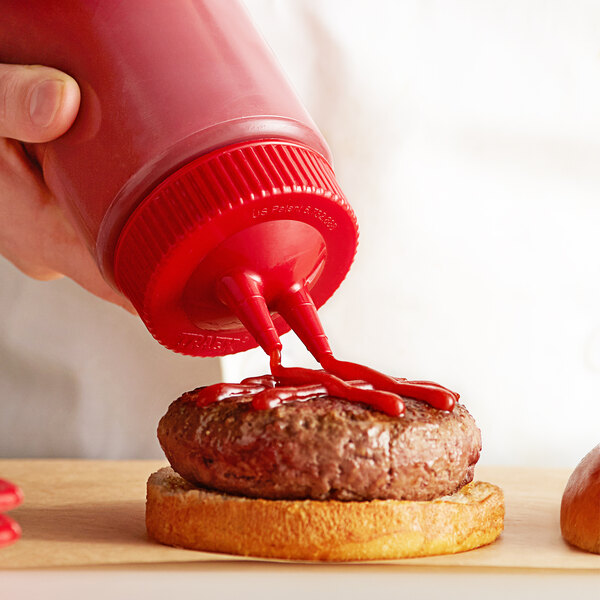 A hand using a Vollrath Twin Tip squeeze bottle with red cap to pour ketchup onto a hamburger.