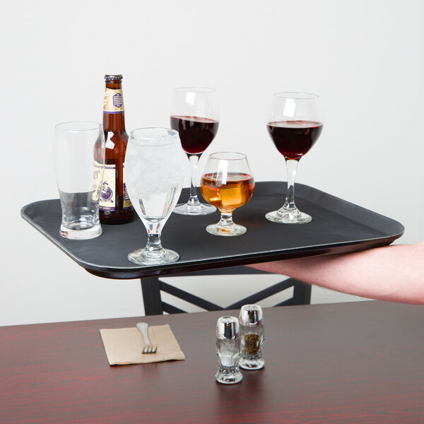 A person holding a Carlisle black non-skid fiberglass serving tray with wine glasses and bottles of alcohol on it.