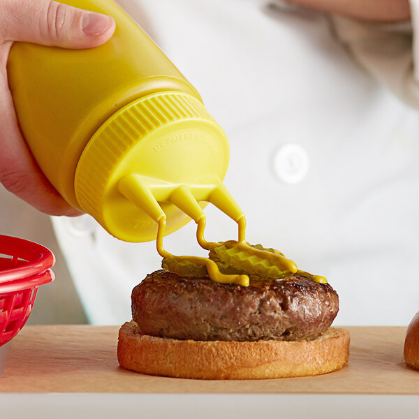 A hand using a Vollrath yellow Tri Tip squeeze bottle to put mustard on a burger.