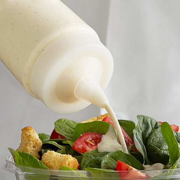 A salad with tomatoes, croutons, and dressing being poured into a bowl using a Vollrath clear wide mouth squeeze bottle.