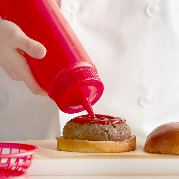 A hand using a Vollrath red wide mouth squeeze bottle to pour ketchup on a hamburger.